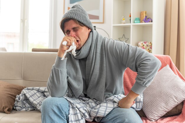 jovem doente com um cachecol no pescoço e chapéu de inverno, espirra segurando a mão perto do rosto, sentado no sofá da sala