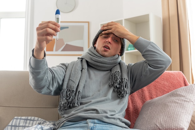 jovem doente com um lenço no pescoço e um chapéu de inverno, colocando a mão na testa e segurando um termômetro sentado no sofá da sala