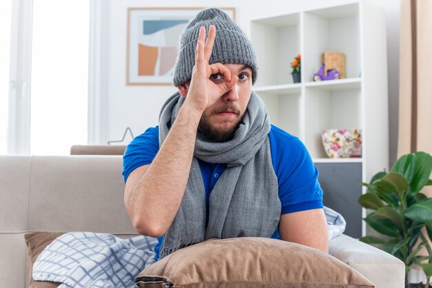 Jovem doente impressionado com cachecol e chapéu de inverno, sentado no sofá na sala de estar com uma almofada nas pernas, olhando para a câmera, fazendo um gesto de olhar