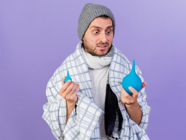 Jovem doente preocupado com chapéu de inverno com lenço segurando e olhando para enema isolado no fundo roxo