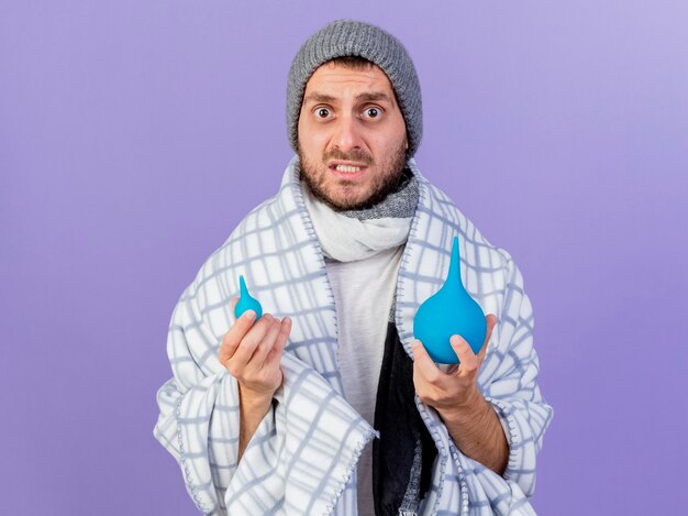 Jovem doente preocupado com chapéu de inverno e lenço segurando enemas isolados no fundo roxo