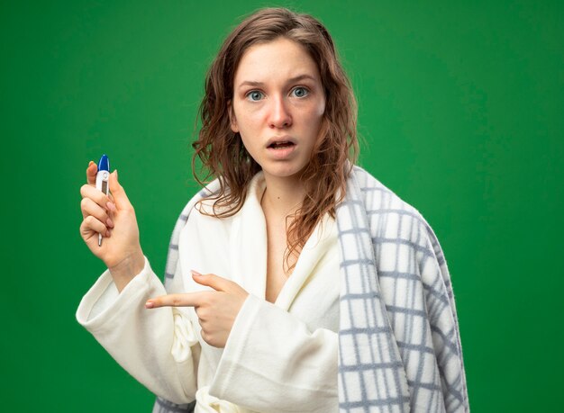 Jovem doente surpreendida com um manto branco embrulhado em xadrez segurando e aponta para um termômetro isolado em verde