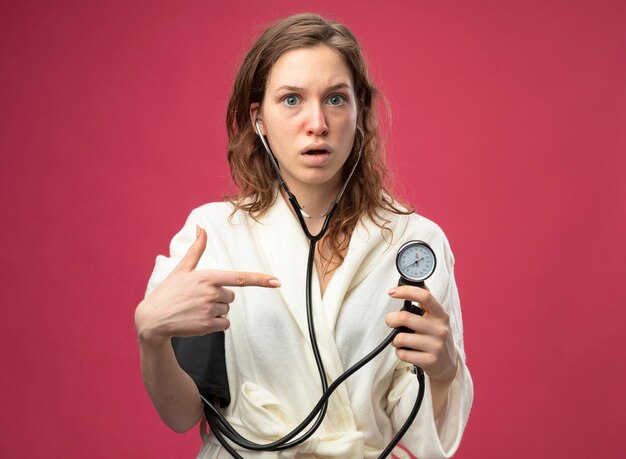 Jovem doente surpreendida olhando para a frente usando um manto branco medindo sua própria pressão com esfigmomanômetro isolado em rosa