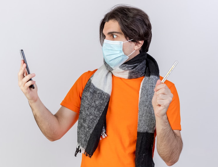 Foto grátis jovem doente surpreso com cachecol e máscara médica, segurando um termômetro, olhando para o telefone na mão, isolado no fundo branco