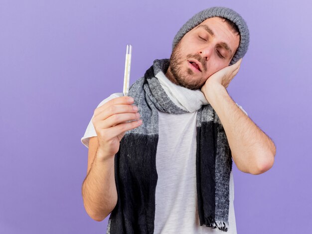 jovem doente usando chapéu de inverno com lenço segurando o termômetro e colocando a mão na bochecha isolada no roxo