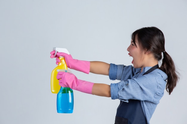 Jovem dona de casa está usando luvas amarelas durante a limpeza com o produto de limpeza na parede branca.