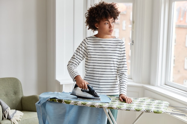 Foto grátis jovem dona de casa pensativa, de pele escura, vestindo roupas listradas, passa roupas na tábua de passar roupa, usa suportes elétricos de ferro e olha pela janela