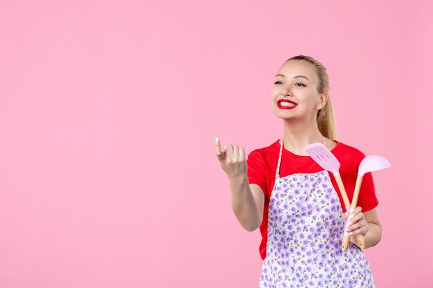 Jovem dona de casa segurando talheres e chamando alguém na parede rosa de frente