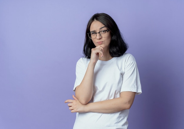 Jovem, duvidosa, bonita, caucasiana, usando óculos, em pé, com a postura fechada, mantendo a mão no queixo isolada em um fundo roxo com espaço de cópia