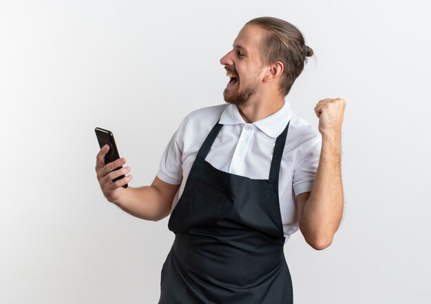 Jovem e alegre barbeiro bonito vestindo uniforme, segurando um telefone celular e levantando o punho com os olhos fechados, isolado no branco