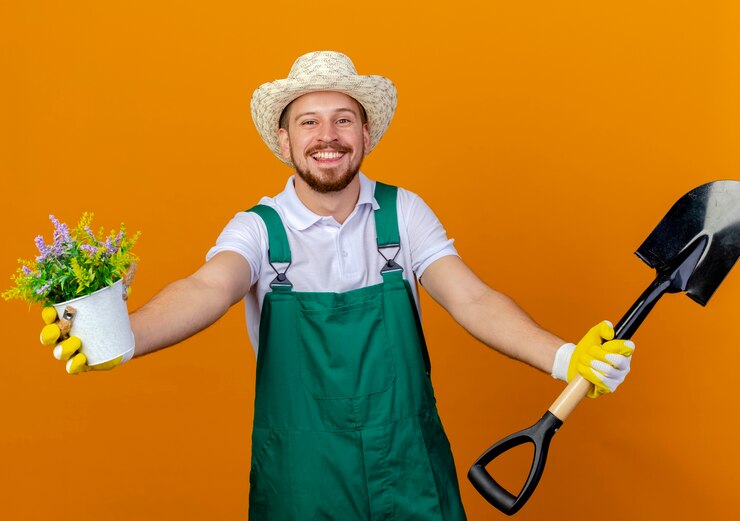 Foto grátis jovem e alegre jardineiro eslavo bonito de uniforme, usando chapéu e luvas de jardinagem, estendendo a pá e o vaso de flores isolados