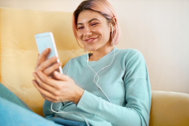 Jovem e alegre mulher europeia com piercing facial relaxando em casa com o telefone inteligente