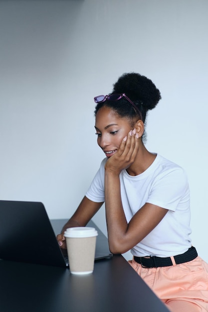 Jovem e atraente afro-americana casual trabalhando alegremente em laptop com café para ir ao escritório moderno