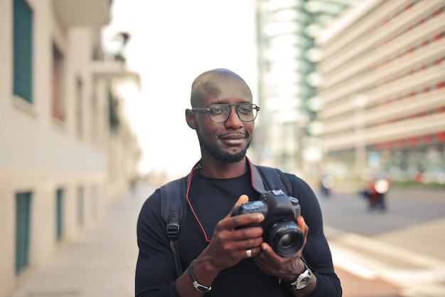 Jovem e atraente fotógrafo africano do sexo masculino com uma câmera em uma rua sob o sol