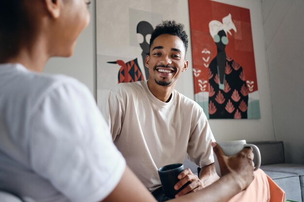 Jovem e atraente homem afro-americano casual sorridente olhando alegremente para a câmera durante a pausa para o café no espaço de trabalho moderno