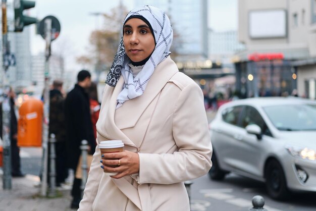 Jovem e atraente mulher árabe em hijab sonhadoramente andando pela rua da cidade com café para ir