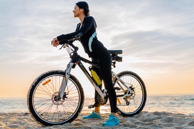 Jovem e atraente mulher magra andando de bicicleta, esporte na praia de verão ao nascer do sol da manhã em roupas de ginástica esportiva, estilo de vida saudável e ativo, sorrindo, feliz se divertindo