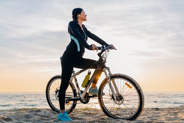Jovem e atraente mulher magra andando de bicicleta, esporte na praia de verão ao nascer do sol da manhã em roupas de ginástica esportiva, estilo de vida saudável e ativo, sorrindo, feliz se divertindo