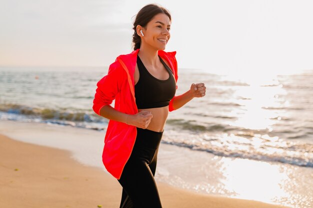 Jovem e atraente mulher magra fazendo exercícios ao nascer do sol da manhã, correndo na praia do mar com roupas esportivas, estilo de vida saudável, ouvindo música nos fones de ouvido e vestindo jaqueta corta-vento rosa