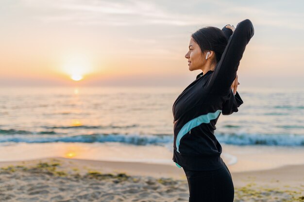 Jovem e atraente mulher magra fazendo exercícios de esporte na praia ao nascer do sol de manhã em roupas esportivas, estilo de vida saudável, ouvindo música em fones de ouvido, fazendo alongamento