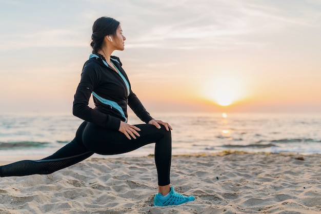 Jovem e atraente mulher magra fazendo exercícios de esporte na praia ao nascer do sol de manhã em roupas esportivas, estilo de vida saudável, ouvindo música em fones de ouvido, fazendo alongamento