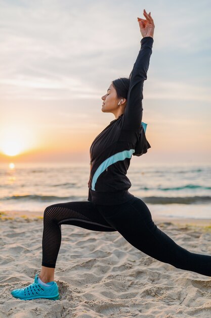 Jovem e atraente mulher magra fazendo exercícios de esporte na praia ao nascer do sol de manhã em roupas esportivas, estilo de vida saudável, ouvindo música em fones de ouvido, fazendo alongamento