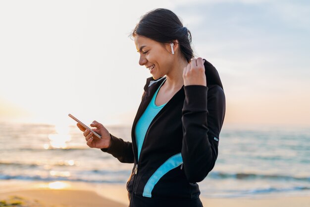 Jovem e atraente mulher magra fazendo exercícios de esporte na praia ao nascer do sol de manhã em roupas esportivas, estilo de vida saudável, ouvindo música em fones de ouvido sem fio segurando um smartphone, sorrindo feliz se divertindo