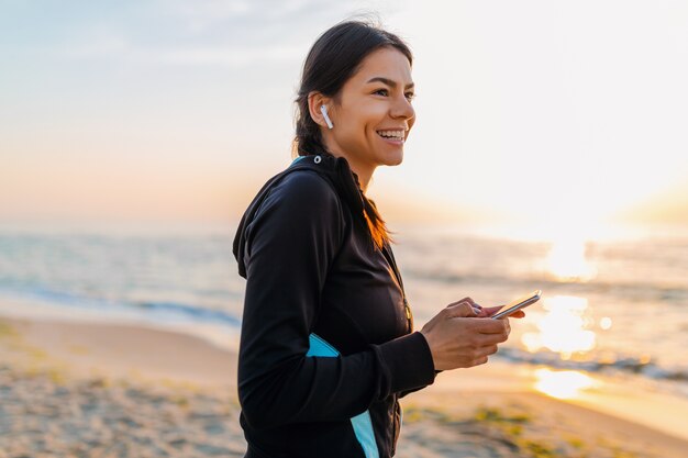 Jovem e atraente mulher magra fazendo exercícios de esporte na praia ao nascer do sol de manhã em roupas esportivas, estilo de vida saudável, ouvindo música em fones de ouvido sem fio segurando um smartphone, sorrindo feliz