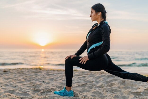 Jovem e atraente mulher magra fazendo exercícios esportivos na praia ao nascer do sol de manhã em roupas esportivas, estilo de vida saudável, ouvindo música em fones de ouvido, fazendo alongamento para as pernas