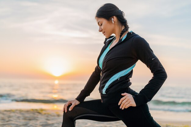 Jovem e atraente mulher magra fazendo exercícios esportivos na praia ao nascer do sol de manhã em roupas esportivas, estilo de vida saudável, ouvindo música em fones de ouvido, fazendo alongamento para as pernas