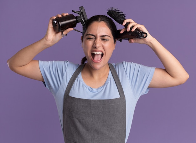 Jovem e bela cabeleireira com avental segurando uma máquina de aparar borrifador e cabelo gritando com uma escova de expressão irritada em pé sobre a parede roxa