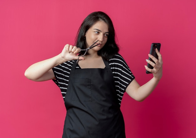 Jovem e bela cabeleireira de avental olhando para a tela do smartphone na chamada online, mostrando uma tesoura na mão sobre a parede rosa