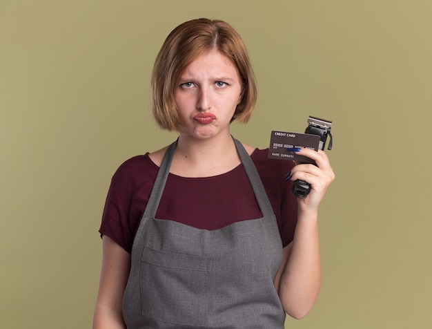 Foto grátis jovem e bela cabeleireira de avental segurando o aparador e o cartão de crédito, olhando para a frente com uma expressão triste em pé sobre a parede verde