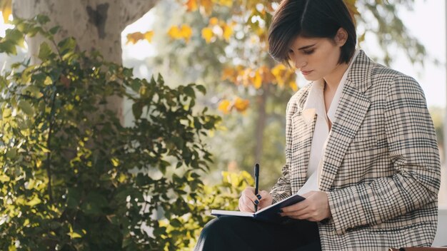 Jovem e bela empresária vestida de blazer xadrez parecendo confiante sentada na rua e fazendo anotações em seu caderno Senhora trabalhando ao ar livre
