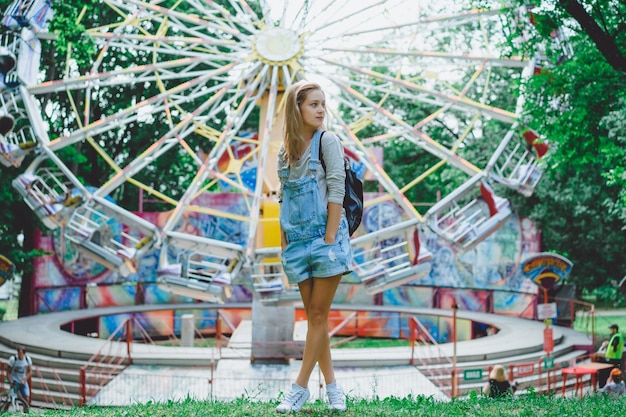 jovem e bela garota loira em denim em geral com uma mochila posando em um parque de diversões