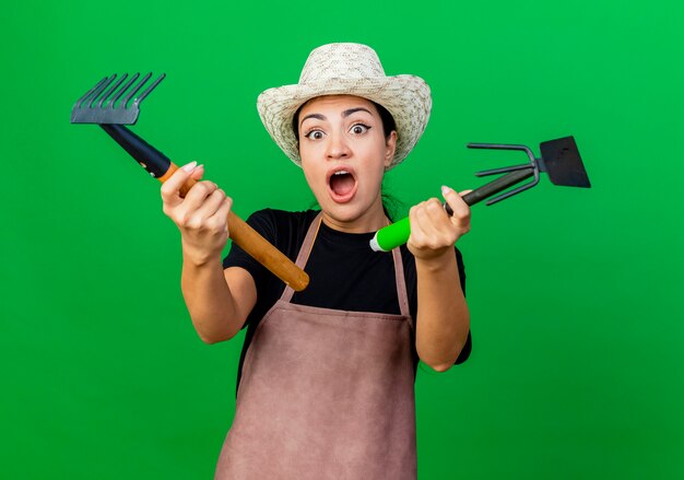 Foto grátis jovem e bela jardineira de avental e chapéu segurando equipamentos de jardinagem, gritando e sendo surpreendida