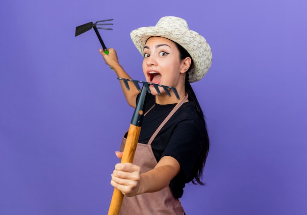 Jovem e bela jardineira de avental e chapéu segurando um equipamento de jardinagem, gritando e sendo surpreendida em pé sobre a parede azul