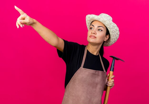 Jovem e bela jardineira de avental e chapéu segurando um mini ancinho apontando para algo com o dedo indicador em pé sobre a parede rosa