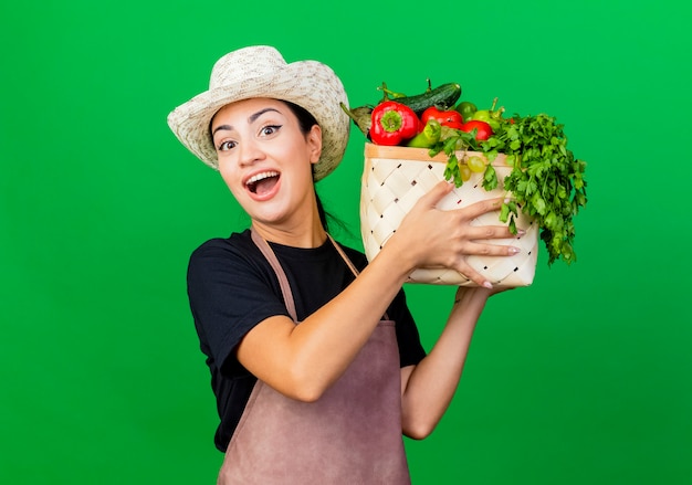 Foto grátis jovem e bela jardineira de avental e chapéu segurando uma caixa cheia de legumes, olhando para a frente, sorrindo com uma cara feliz em pé sobre a parede verde