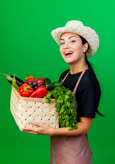 Jovem e bela jardineira de avental e chapéu segurando uma cesta cheia de vegetais e sorrindo com uma cara feliz