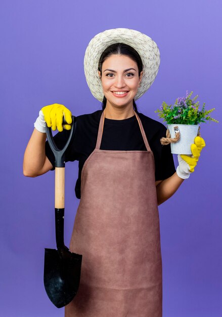 Jovem e bela jardineira de avental e chapéu segurando uma pá e um vaso de plantas olhando para frente com um sorriso no rosto em pé sobre a parede azul