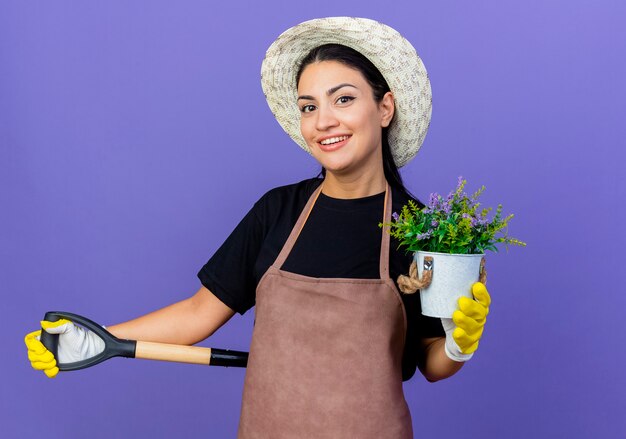 Jovem e bela jardineira de avental e chapéu segurando uma pá, mostrando um vaso de planta sorrindo alegremente em pé sobre a parede azul