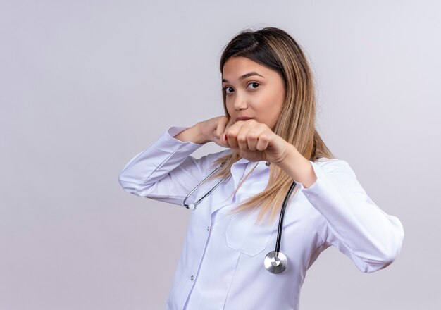 Jovem e bela médica vestindo jaleco branco com estetoscópio, cara séria, posando de boxeador com os punhos cerrados