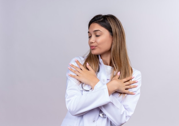 Jovem e bela médica vestindo jaleco branco com estetoscópio em pé com as mãos cruzadas no peito sentindo emoções positivas