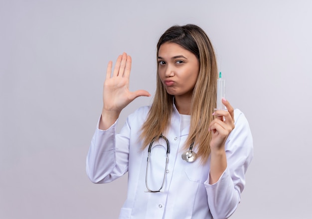 Jovem e bela médica vestindo jaleco branco com estetoscópio segurando uma seringa parecendo descontente em pé com a mão aberta fazendo sinal de pare