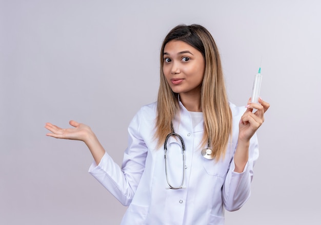Jovem e bela médica vestindo jaleco branco com estetoscópio segurando uma seringa