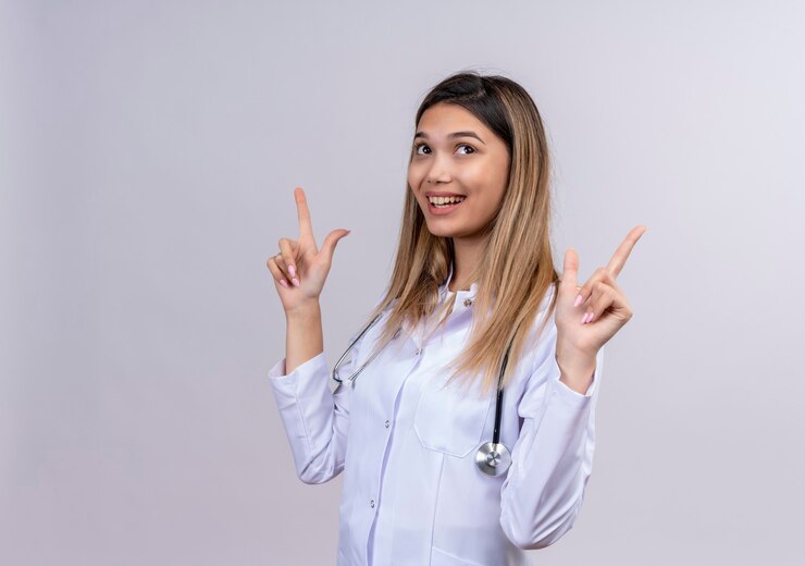 Foto grátis jovem e bela médica vestindo jaleco branco com estetoscópio sorrindo alegremente apontando o dedo indicador para cima