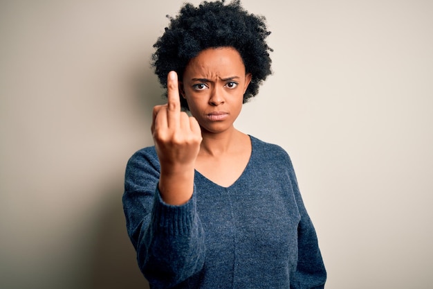 Jovem e bela mulher afro-americana com cabelos cacheados, vestindo um suéter casual, mostrando o dedo médio, indelicado e rude, foda-se a expressão