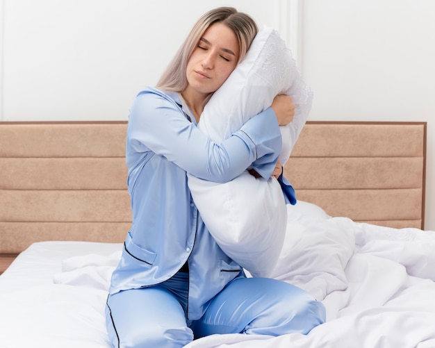 Foto grátis jovem e bela mulher de pijama azul, sentada na cama, abraçando o travesseiro com os olhos fechados, sentindo emoções positivas no interior do quarto com luz de fundo
