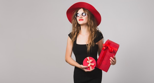 Jovem e bela mulher segurando presentes, vestido preto, chapéu vermelho, óculos de sol, feliz, sorridente, sexy, elegante, caixas de presente, comemorando, positiva